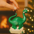 Load image into Gallery viewer, Green dinosaur ornament held by a hand with a warm background
