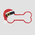 Load image into Gallery viewer, Personalized dog Christmas ornament featuring a bone shaped design with a glitter red Santa hat detail on one end, customizable name area centered on the ornament and customizable year displayed on the opposite side, smooth glossy finish with glitter accents, flat lightweight structure and red hanging ribbon, photographed on a snowy holiday styled background with pine branches, festive Christmas ornaments, dog themed props, and warm seasonal lighting for pet lovers.
