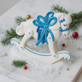 Load image into Gallery viewer, White rocking horse ornament with a blue bow on a snowy background with Christmas decorations.
