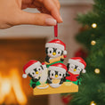 Load image into Gallery viewer, Christmas ornament with penguins in red hats and green scarves against a blurred Christmas tree background.
