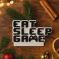Load image into Gallery viewer, Personalized Eat Sleep Game Christmas ornament with a raised 3D front featuring bold block-style lettering and a detailed game controller icon. The front design is fully three dimensional, creating depth and texture, while the back is flat 2D for easy hanging and durability. White base with black raised letters and subtle colored accents. Includes hand-added personalization with name and year or family name. Gaming-themed holiday ornament displayed on snowy festive background with warm lights, designed for 
