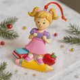 Load image into Gallery viewer, Decorative Christmas ornament of a girl with a pencil, apple, and books on a snowy background.
