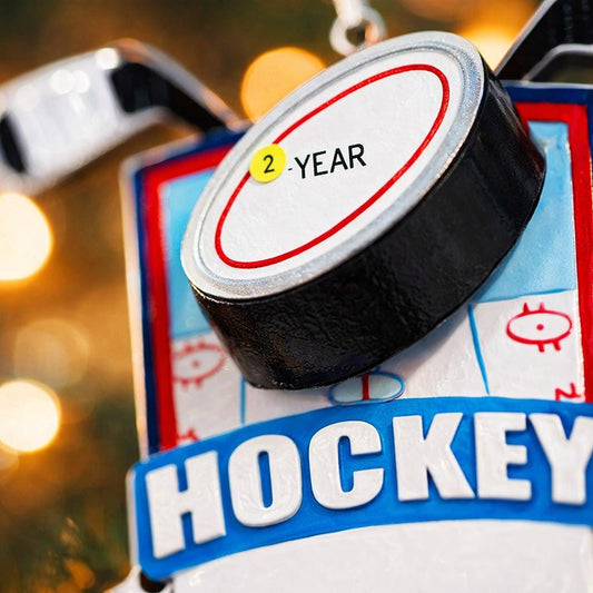 Personalized hockey Christmas ornament featuring a hockey puck and crossed hockey sticks design, customizable name area on the front banner and customizable year area on the puck, glossy finish with raised 3D front details and flat 2D back, white hanging ribbon, displayed on a snowy ice rink themed holiday background with hockey accessories and festive winter decorations.