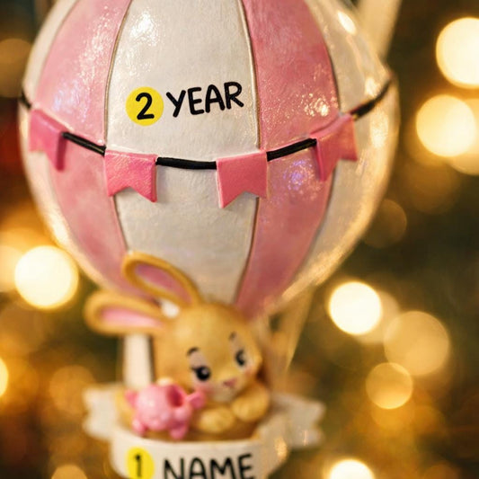 A personalized baby girl ornament featuring a cute bunny riding a pastel pink hot air balloon. Soft pink and white tones with delicate bow details create a sweet and cozy look. Handwritten personalization includes baby name and year. Mostly 2D ornament with raised 3D details on the front only, lightweight and easy to hang, perfect for baby first Christmas, nursery decor, and toddler keepsakes.