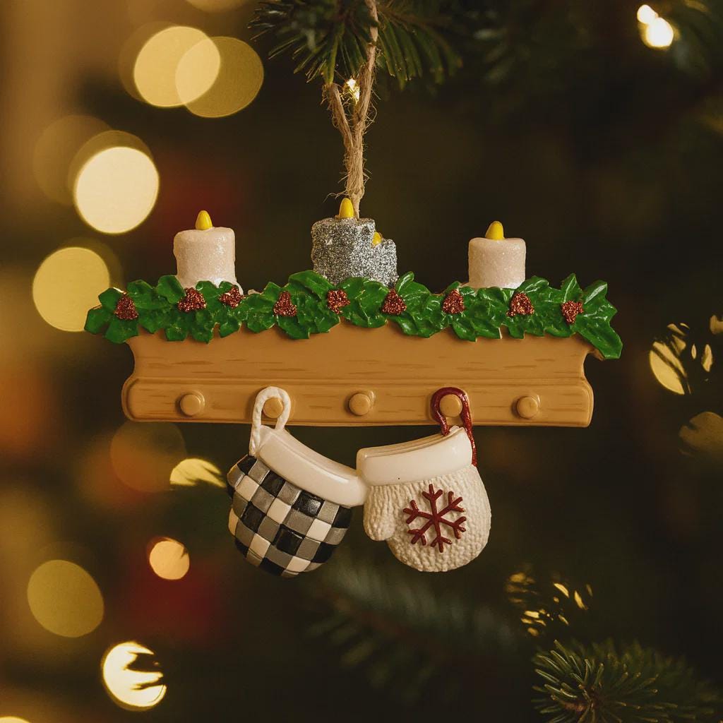 Christmas ornament with teapot, mittens, and candles on a wooden shelf against a blurred Christmas tree background.