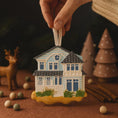 Load image into Gallery viewer, Decorative house ornament being held above a festive scene with trees and deer.
