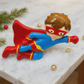 Load image into Gallery viewer, Toy ornament of a superhero in a red and blue costume with a cape on a marble surface.

