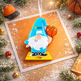 Load image into Gallery viewer, Personalized basketball gnome Christmas ornament featuring a cute blue gnome character holding a basketball, customizable name area on the hat and customizable year area on the base, glossy finish with raised 3D front details and flat 2D back, white hanging ribbon, displayed on a snowy holiday background with pine branches and festive decorations.

