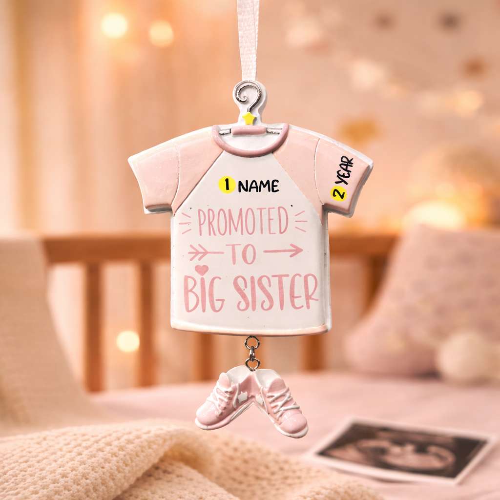 Decorative ornament shaped like a baby shirt with 'Promoted to Big Sister' text, hanging in a softly blurred room.