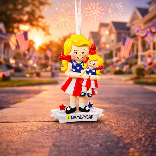 Decorative ornament of a woman and child in American flag outfits with a street scene and fireworks in the background.