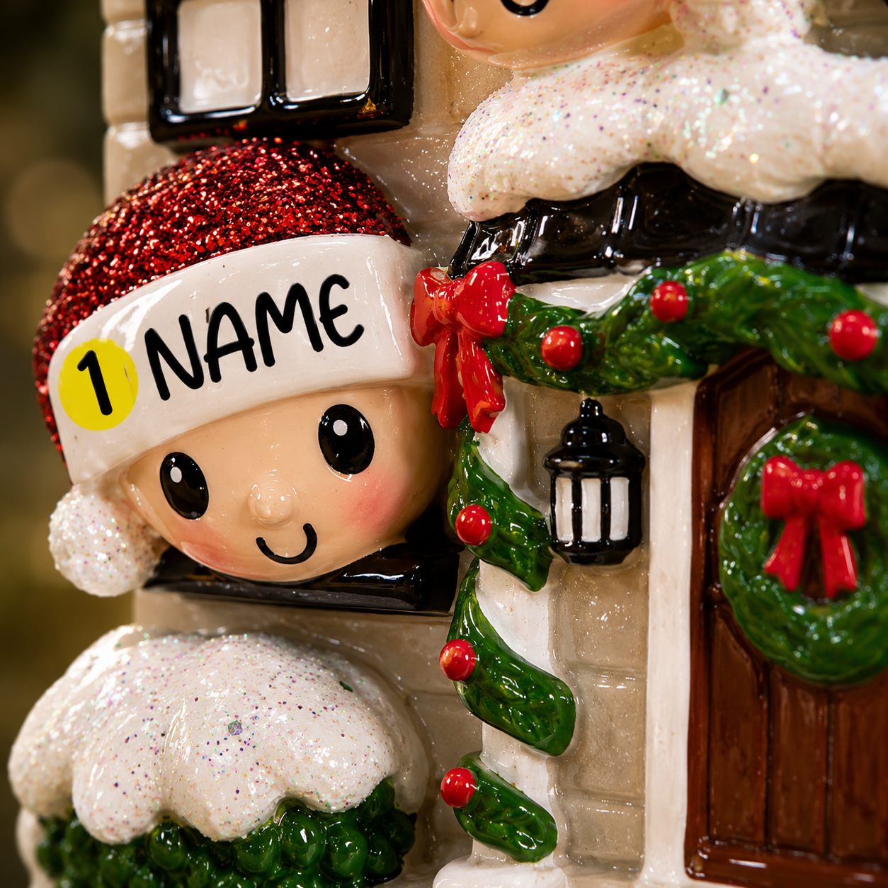 Decorative Christmas ornament with a child wearing a red hat and 'NAME' label, standing in front of a house with wreaths and a lantern.