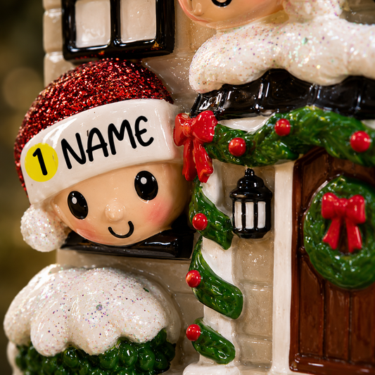 Decorative Christmas ornament with a child wearing a red hat and 'NAME' label, standing in front of a house with wreaths and a lantern.