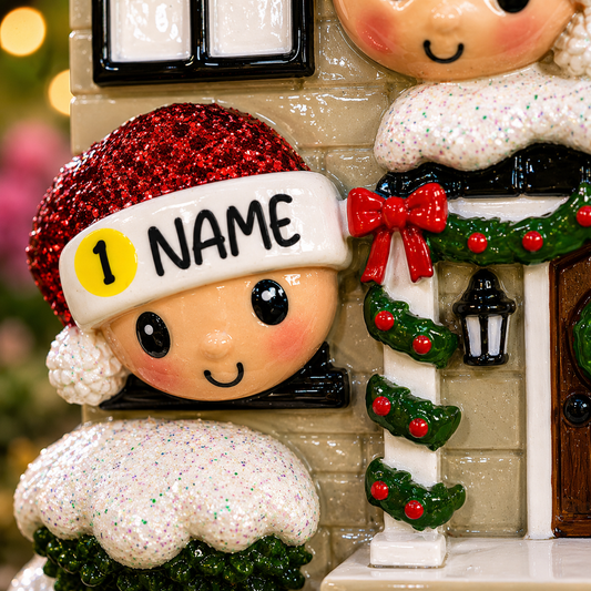 Decorative Christmas ornament with a child wearing a red hat and 'NAME' headband, standing next to a house with a wreath.