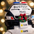 Load image into Gallery viewer, Personalized police car Christmas ornament featuring a black and white cop car design with BUSTED text, police lights and festive wreath detail. Customized with a name and year on the front banner. Glossy resin finish with bold black and white colors creates a fun and playful holiday look. Mostly 2D ornament with raised 3D details on the front only, lightweight and perfect for Christmas tree decor and police themed gifts.
