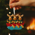Load image into Gallery viewer, Decorative ornament of three moose in a canoe with a blurred Christmas tree and fireplace in the background.
