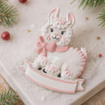 Load image into Gallery viewer, A glossy resin baby girl llama ornament features a white smiling llama with soft pink accents, raised textured fur details, pink bow and scarf elements, glitter highlights, and a smooth painted cartoon style finish, styled with warm holiday lights and nursery themed props, handwritten personalization showing the baby name on the front banner and the year placed on the llama body, designed as a mostly 2D ornament with raised 3D details on the front only.
