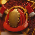 Load image into Gallery viewer, Close-up of a red and gold helmet with a blurred red glittery background
