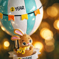 Load image into Gallery viewer, A personalized baby boy ornament featuring a cute bunny riding a pastel blue hot air balloon. Soft blue and white tones with gentle decorative details create a calm and cozy look. Handwritten personalization includes baby name and year. Mostly 2D ornament with raised 3D details on the front only, lightweight and easy to hang, perfect for baby first Christmas, nursery decor, and toddler keepsakes.
