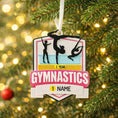 Load image into Gallery viewer, Personalized gymnastics Christmas ornament featuring elegant gymnast silhouette figures in motion, customizable name area on the front banner and customizable year area above the banner, glossy finish with raised 3D front details and flat 2D back, white hanging ribbon, displayed on a soft pink snowy holiday background with festive ornaments greenery and warm Christmas decor.

