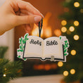 Load image into Gallery viewer, Personalized Holy Bible Christmas Ornament featuring an open scripture design with elegant “Holy Bible” lettering across the pages. The left page is customized with a name and the right page displays the year, creating a meaningful faith based keepsake. Surrounded by soft greenery and delicate flowers, this religious ornament is perfect for baptisms, baby dedications, first communions, or celebrating a Christ centered Christmas. A thoughtful spiritual gift for families who cherish their faith.
