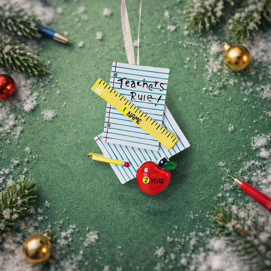 Decorative ornament shaped like paper book with a ruler, pen, and red apple on a green background with Christmas elements.