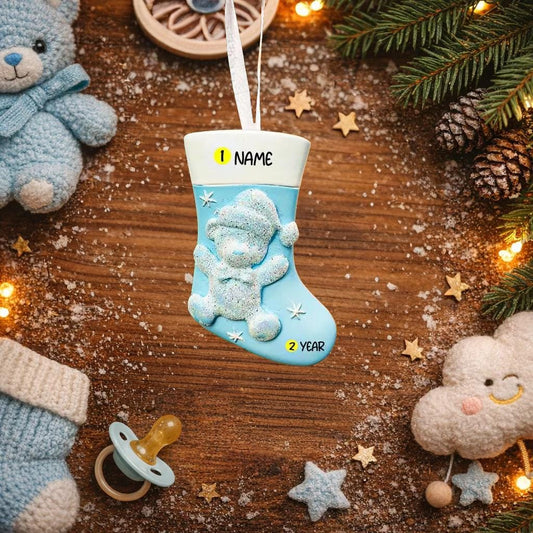 A glossy resin baby boy stocking ornament features a soft pastel blue Christmas stocking shape with a raised glitter style baby figure on the front, white cuff detail, small star accents, and smooth painted surfaces, surrounded by warm holiday lights and nursery themed props, handwritten personalization showing one baby name on the top cuff and the year written near the bottom of the stocking, designed as a mostly 2D ornament with raised 3D details on the front only.