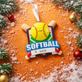 Load image into Gallery viewer, Personalized softball Christmas ornament featuring a yellow softball with stitched details and crossed bats design on a shield style background, customizable name area on the front banner and customizable year area on the softball, glossy finish with raised 3D front details and flat 2D back, white hanging ribbon, displayed on a snowy softball field themed holiday background with pine branches and festive decorations.
