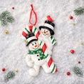 Load image into Gallery viewer, Two snowman ornaments holding a candy cane on a snowy background with Christmas decorations.
