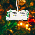 Load image into Gallery viewer, Personalized Holy Bible Christmas Ornament featuring an open scripture design with elegant “Holy Bible” lettering across the pages. The left page is customized with a name and the right page displays the year, creating a meaningful faith based keepsake. Surrounded by soft greenery and delicate flowers, this religious ornament is perfect for baptisms, baby dedications, first communions, or celebrating a Christ centered Christmas. A thoughtful spiritual gift for families who cherish their faith.
