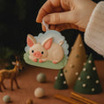 Load image into Gallery viewer, Hand holding a pig-shaped ornament with a forest background
