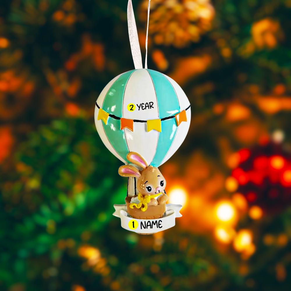 A personalized baby boy ornament featuring a cute bunny riding a pastel blue hot air balloon. Soft blue and white tones with gentle decorative details create a calm and cozy look. Handwritten personalization includes baby name and year. Mostly 2D ornament with raised 3D details on the front only, lightweight and easy to hang, perfect for baby first Christmas, nursery decor, and toddler keepsakes.