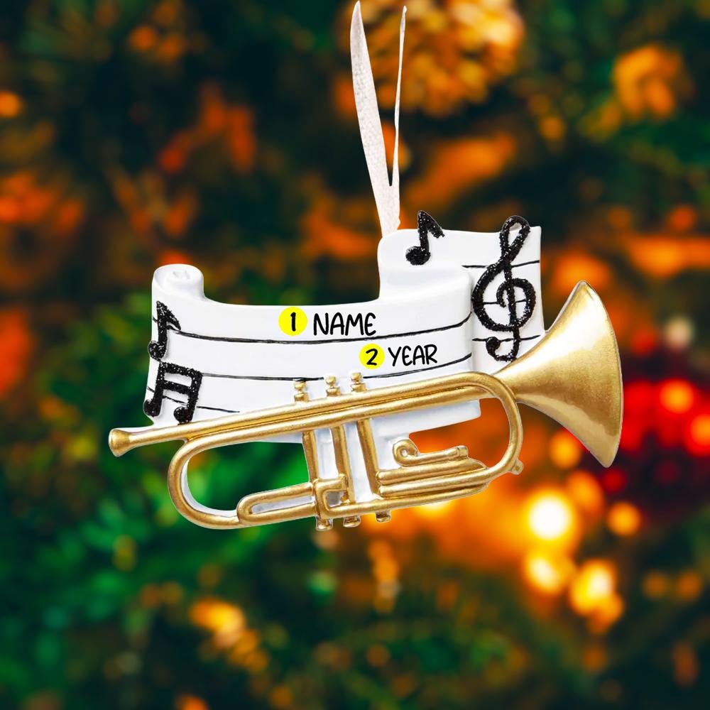 Personalized music ornament featuring a white musical staff design with black notes and a gold trumpet detail. Includes free handwritten personalization with one name and year written on the sheet music. Shown hanging with a ribbon in a warm festive holiday setting with soft lights instruments and seasonal decor. Designed with a 3D raised front and flat back perfect for Christmas tree display and gifts for musicians music lovers and band members.