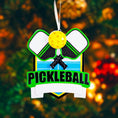 Load image into Gallery viewer, Personalized pickleball Christmas ornament featuring crossed pickleball paddles and a yellow pickleball on a shield style background, customizable name area on the front banner and customizable year area on the ball, glossy finish with raised 3D front details and flat 2D back, white hanging ribbon, displayed on a snowy pickleball court themed holiday background with pine branches and festive decorations.

