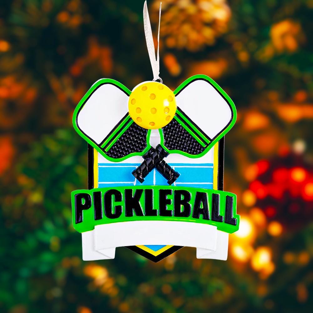 Personalized pickleball Christmas ornament featuring crossed pickleball paddles and a yellow pickleball on a shield style background, customizable name area on the front banner and customizable year area on the ball, glossy finish with raised 3D front details and flat 2D back, white hanging ribbon, displayed on a snowy pickleball court themed holiday background with pine branches and festive decorations.