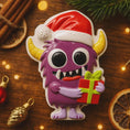 Load image into Gallery viewer, Purple monster character with a Santa hat holding a gift on a wooden surface with Christmas decorations.
