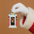 Load image into Gallery viewer, Hand wearing a white glove holding a small decorative house ornament with a wreath on a brown background
