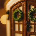 Load image into Gallery viewer, Close-up of a gingerbread house door with wreaths and a blurred light in the background
