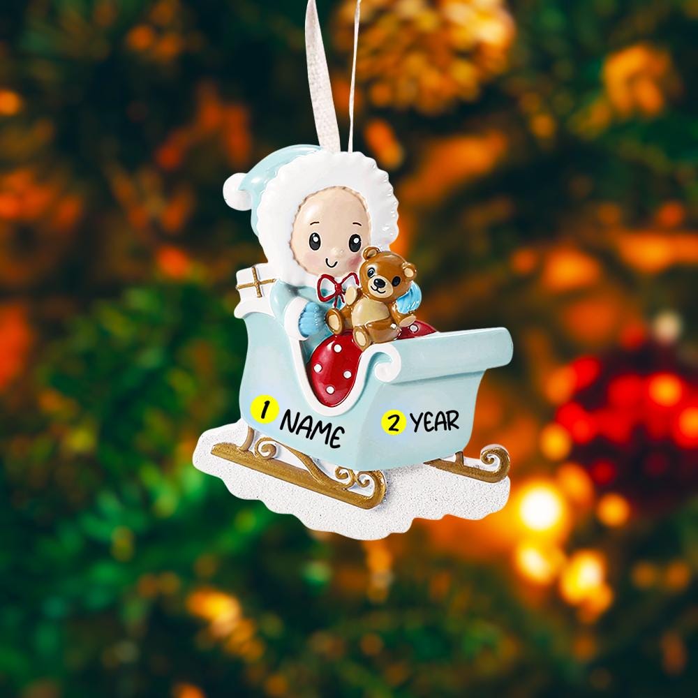 Personalized baby first Christmas ornament featuring a sweet baby boy sitting in a pastel blue sleigh holding a small teddy bear. Handwritten baby name and year displayed on the sleigh front. Smooth resin finish with soft winter tones, lightweight structure, mostly 2D ornament with raised 3D front details, designed for baby boy nursery themed Christmas tree decor and keepsake gifting.
