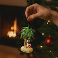 Load image into Gallery viewer, Decorative palm tree ornament with lights and a gift, held by a hand in front of a Christmas tree and fireplace.
