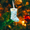Load image into Gallery viewer, A glossy resin baby boy stocking ornament features a soft pastel blue Christmas stocking shape with a raised glitter style baby figure on the front, white cuff detail, small star accents, and smooth painted surfaces, surrounded by warm holiday lights and nursery themed props, handwritten personalization showing one baby name on the top cuff and the year written near the bottom of the stocking, designed as a mostly 2D ornament with raised 3D details on the front only.
