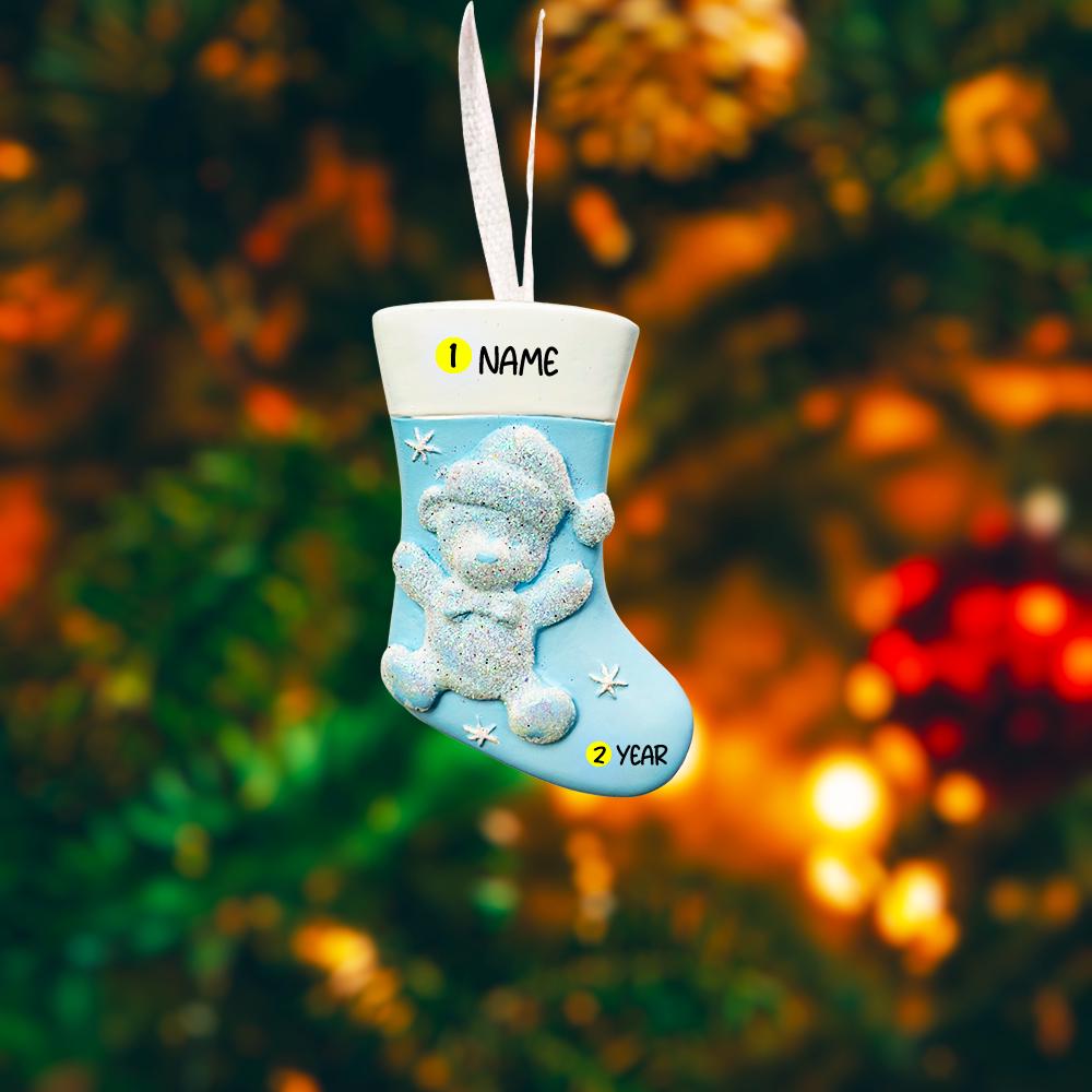 A glossy resin baby boy stocking ornament features a soft pastel blue Christmas stocking shape with a raised glitter style baby figure on the front, white cuff detail, small star accents, and smooth painted surfaces, surrounded by warm holiday lights and nursery themed props, handwritten personalization showing one baby name on the top cuff and the year written near the bottom of the stocking, designed as a mostly 2D ornament with raised 3D details on the front only.