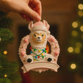 Load image into Gallery viewer, Personalized pink llama Christmas ornament featuring a cute white llama wrapped in colorful holiday lights, framed in a glittery wreath with pink accents. The top hat band can be customized with a name, while the bottom banner allows space for year or Baby’s 1st Christmas. Designed in soft pink and festive tones, this resin baby girl Christmas ornament makes a meaningful keepsake gift for celebrating a newborn’s first holiday season.

