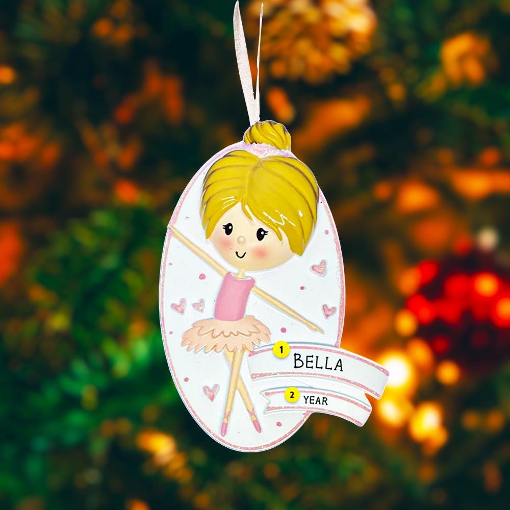 Personalized ballet girl Christmas ornament featuring a cute ballerina character in a pink tutu dress with heart details, customizable name area on the front banner and customizable year area below, glossy finish with raised 3D front details and flat 2D back, white hanging ribbon, displayed on a snowy holiday background with pine branches and festive decorations.