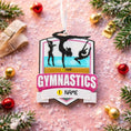 Load image into Gallery viewer, Personalized gymnastics Christmas ornament featuring elegant gymnast silhouette figures in motion, customizable name area on the front banner and customizable year area above the banner, glossy finish with raised 3D front details and flat 2D back, white hanging ribbon, displayed on a soft pink snowy holiday background with festive ornaments greenery and warm Christmas decor.

