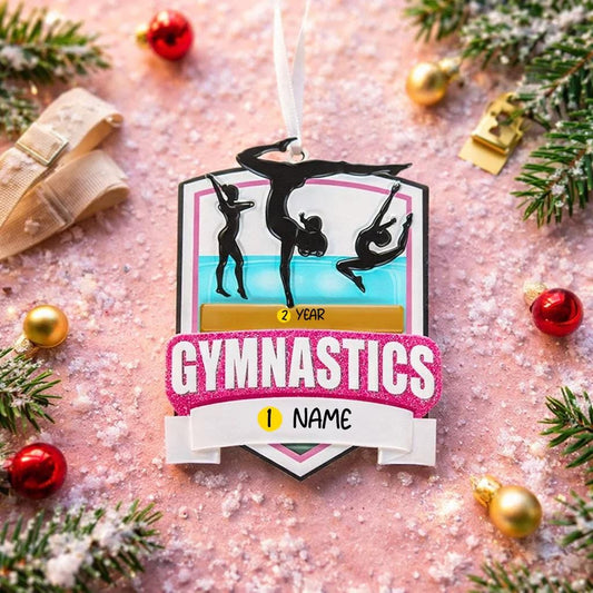 Personalized gymnastics Christmas ornament featuring elegant gymnast silhouette figures in motion, customizable name area on the front banner and customizable year area above the banner, glossy finish with raised 3D front details and flat 2D back, white hanging ribbon, displayed on a soft pink snowy holiday background with festive ornaments greenery and warm Christmas decor.
