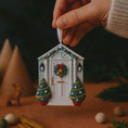 Load image into Gallery viewer, Decorative Christmas house ornament with a wreath and trees, held by a hand against a dark background.
