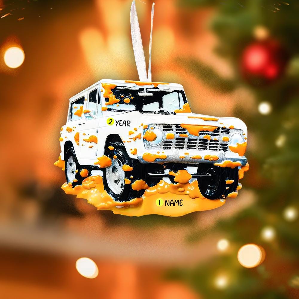 Decorative Christmas ornament of a white truck with orange mud splashes on a blurred Christmas tree background.