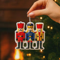 Load image into Gallery viewer, Three nutcracker figurines on a decorative tag held by a hand against a Christmas tree background.

