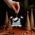 Load image into Gallery viewer, Personalized hockey Christmas ornament featuring a hockey puck and crossed hockey sticks design, customizable name area on the front banner and customizable year area on the puck, glossy finish with raised 3D front details and flat 2D back, white hanging ribbon, displayed on a snowy ice rink themed holiday background with hockey accessories and festive winter decorations.
