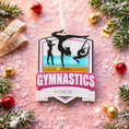 Load image into Gallery viewer, Personalized gymnastics Christmas ornament featuring elegant gymnast silhouette figures in motion, customizable name area on the front banner and customizable year area above the banner, glossy finish with raised 3D front details and flat 2D back, white hanging ribbon, displayed on a soft pink snowy holiday background with festive ornaments greenery and warm Christmas decor.

